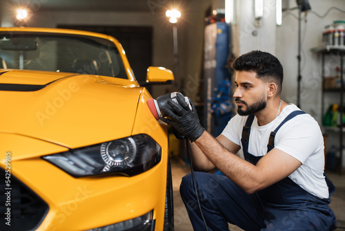 Car detailing and polishing concept. Professional young male car service worker in uniform, holding in hands orbital polisher, and polishing yellow luxury car at auto repair service station.