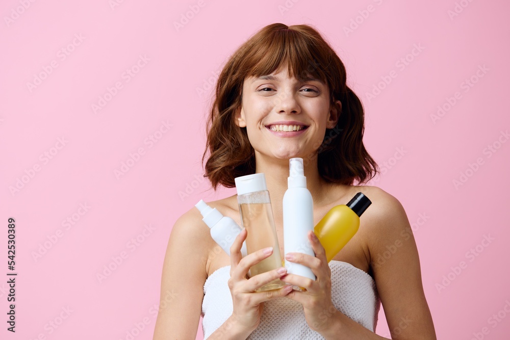 a young, well-groomed woman stands on a pink background holding different jars of care cosmetics in her hands and smiling broadly with her teeth, squints her eyes. Horizontal photo with an empty space a young, well-groomed woman stands on a pink background holding different jars of care cosmetics in her hands and smiling broadly with her teeth, squints her eyes. Horizontal photo with an empty space