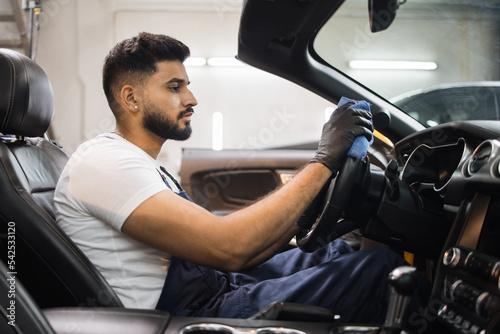 Handsome bearded young man in uniform and protective gloves, cleaning car interior and steering wheel using microfiber cloth, smiling at camera. Car detailing and valeting concept. © sofiko14