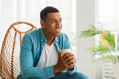 Smiling millennial african american guy with cup of coffee takeaway, sit on chair, enjoy comfort and favorite drink