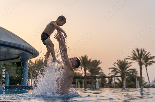 Dad and son are playing in the swimming pool. The father tosses the son up.