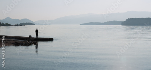 Lone fisherman, in silhouette, on the jetty in early morning mist with hills in the distance