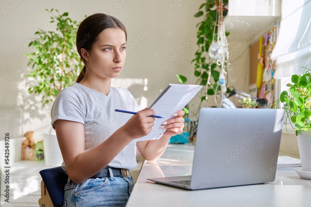 Teenage girl studying at home using laptop making notes in notebook Teenage girl studying at home using laptop making notes in notebook