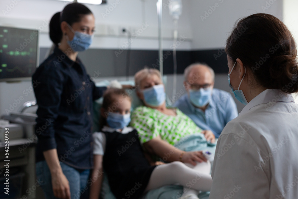 Unrecognizable doctor wearing cover up, visiting room to monitor patient health condition progress. Family wearing medical masks in grandmother hospital bedroom to prevent coronavirus infection. Unrecognizable doctor wearing cover up, visiting room to monitor patient health condition progress. Family wearing medical masks in grandmother hospital bedroom to prevent coronavirus infection.