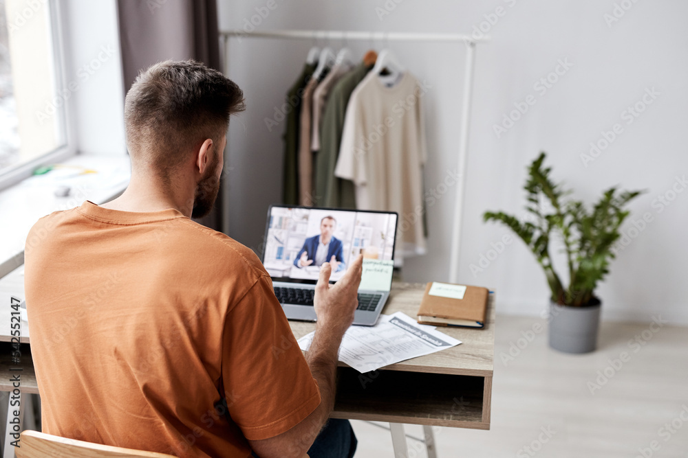 Back view of young man sitting in front of laptop with tutor on screen and asking him questions about new subject points during online lesson Back view of young man sitting in front of laptop with tutor on screen and asking him questions about new subject points during online lesson