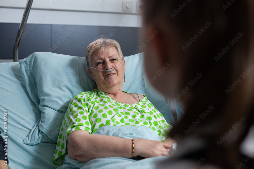 Elderly woman talking to unrecognizable girl. Close up of happy grandmother being visited by granddaughter at geriatric hospital room. Smiling old lady talking to female relative in sanatorium bed. - obrazy, fototapety, plakaty 