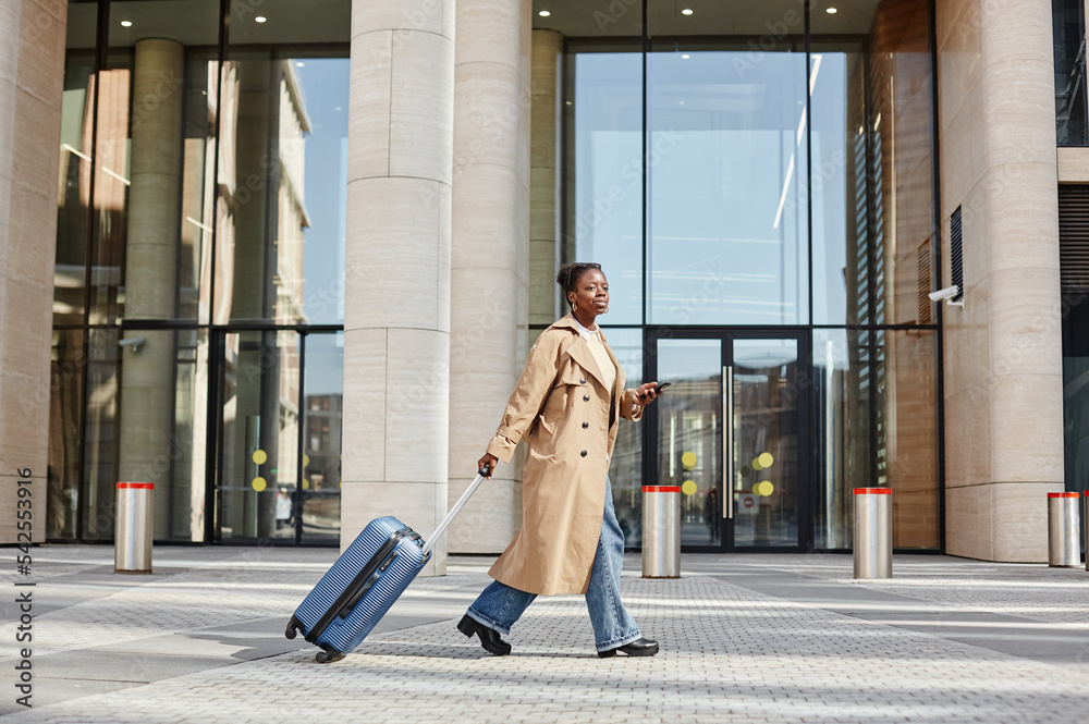 Side view portrait of young black woman with suitcase walking outdoors in city by airport or train station
