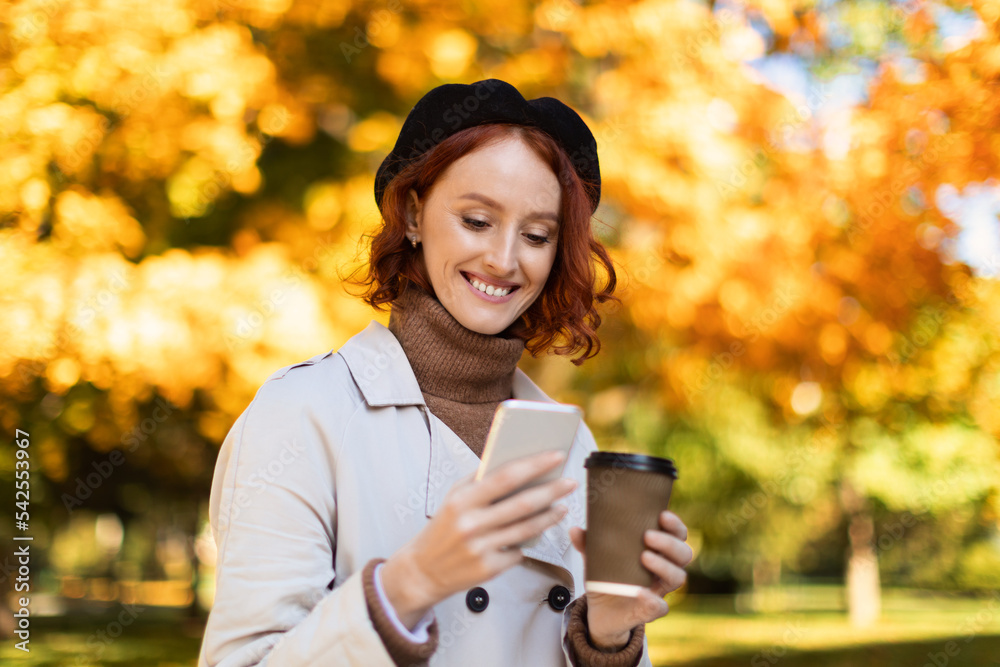 Smiling young european red-haired female in raincoat and had reads message on smartphone, enjoy takeaway drink Smiling young european red-haired female in raincoat and had reads message on smartphone, enjoy takeaway drink
