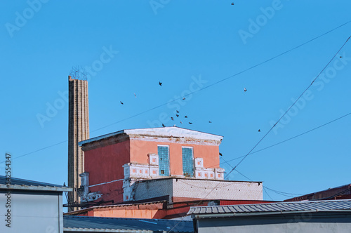 A destroyed or unfinished industrial reinforced concrete chimney next to a production building against the background of a blue sky and a flock of birds. Post-apocalytic landscape