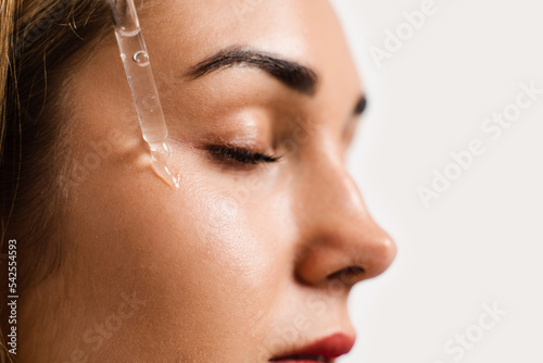 Girl drips facial serum on her cheek for acne treatment and wrinkle smoothing. Girl applies face serum to her face with pipette on white background. photo