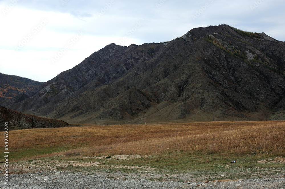 A flat rocky clearing with dried and yellowed grass at the foot of a high mountain.
