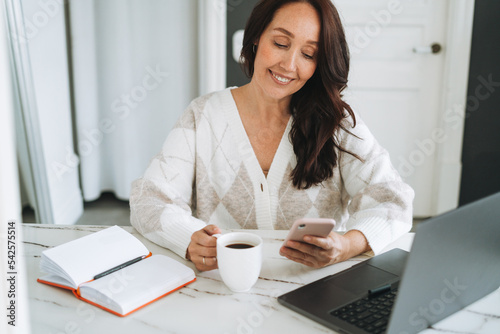 Smiling brunette woman with long hair in white cardigan working on laptop using mobile phone in bright modern office