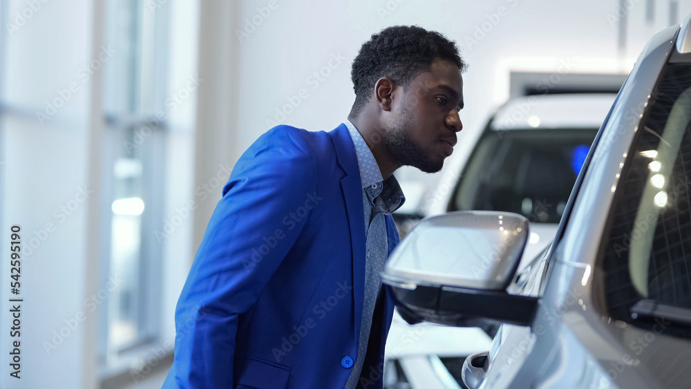 African American man client examines modern car and thinks about purchasing in automobile salon. Black customer tries to search nice vehicle African American man client examines modern car and thinks about purchasing in automobile salon. Black customer tries to search nice vehicle