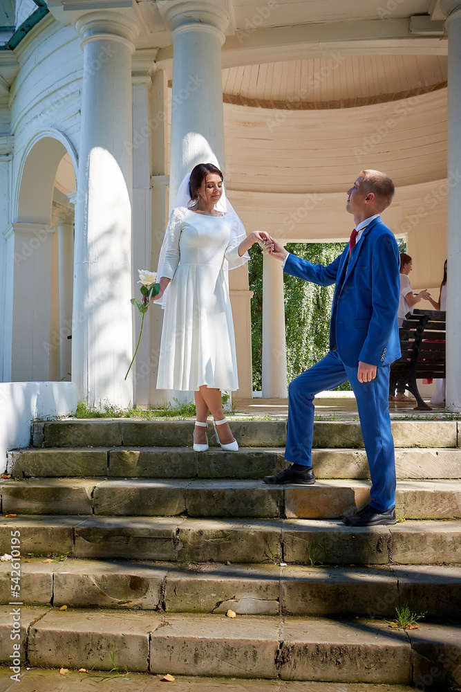 Stylish groom in a blue suit walk with the bride in a white dress in the gazebo near the white columns. Wedding portrait of happy newlyweds in a summer sunny day