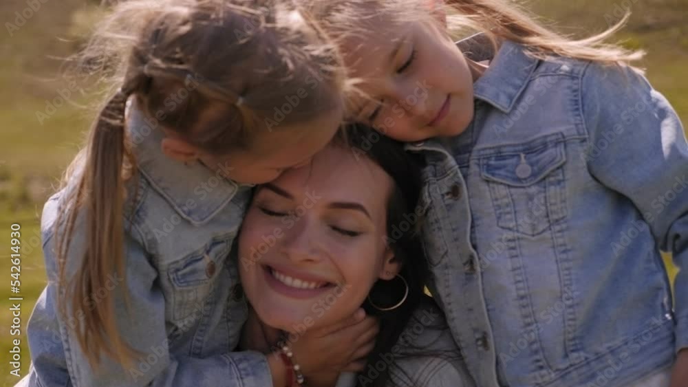 A happy young mother hugs her twin daughters on a field with green grass in Sunny weather. A happy young mother hugs her twin daughters on a field with green grass in Sunny weather.