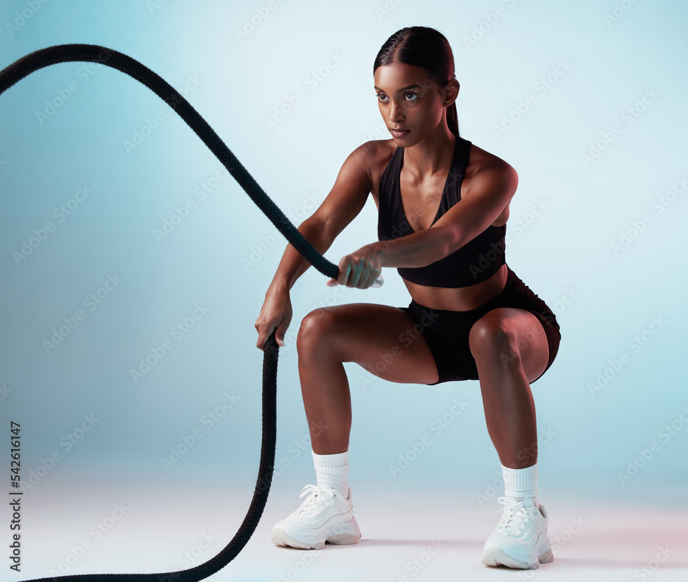 Battle rope, fitness and woman in studio for exercise, strength training  and cardio workout burning calories. Wellness, focus and healthy girl in a  squat pose moving ropes with a blue background Stock