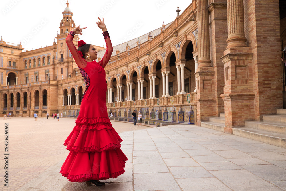 Fototapeta premium Beautiful teenage woman dancing flamenco in a square in Seville, Spain. She wears a red dress with ruffles and dances flamenco with a lot of art. Flamenco cultural heritage of humanity.
