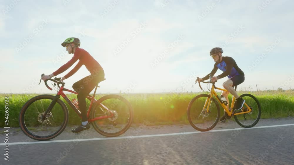 Side view of two male cyclists wearing sport clothes and safety helmet riding bikes on countryside road. Professional sportsman training together outdoors. Active lifestyles concept. Side view of two male cyclists wearing sport clothes and safety helmet riding bikes on countryside road. Professional sportsman training together outdoors. Active lifestyles concept.