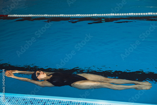 Full length of athletic professional female swimmer showing backstroke while swimming in water of indoor pool during training 