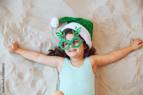 A little girl is playing with sand on the beach in a Santa Claus hat and Christmas glasses. The child is resting on the sea. The concept of Christmas holidays at sea. Space for copying.