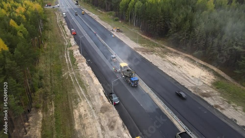 Top view of the work of road machinery. The drone flies over the new asphalt site and descends towards the truck and paver. Workers in overalls are laying hot asphalt. The roller levels road surface.
