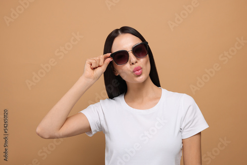 Beautiful young woman in stylish sunglasses blowing kiss on beige background