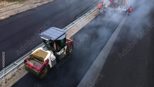 The road roller compacts the hot asphalt after the paver. Thick steam rises over the new road surface. Workers are repairing the road. An empty dump truck goes for a new asphalt mix. Drone footage