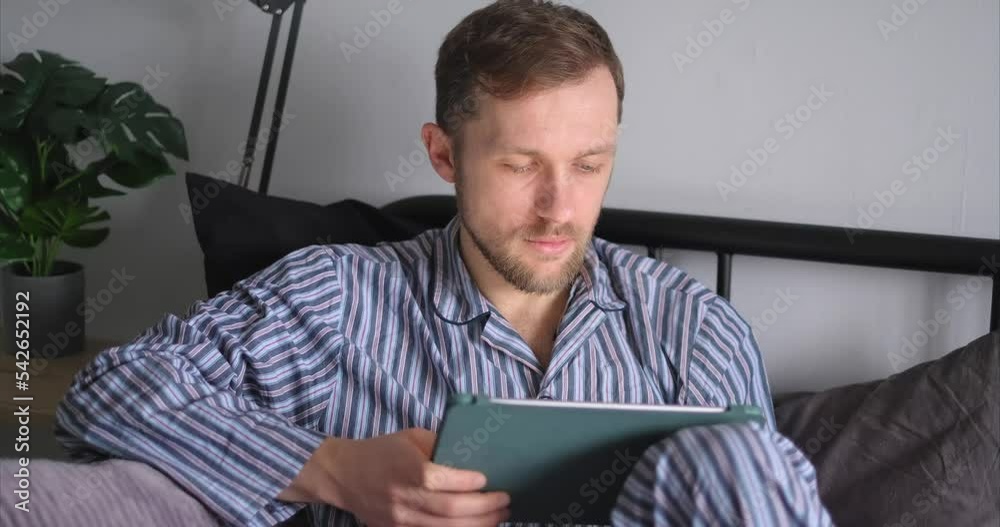 Young cheerful man using tablet computer sitting in bed in the morning making online shopping, working, education or surfing the internet. Caucasian male using digital tablet at home. 4k video footage