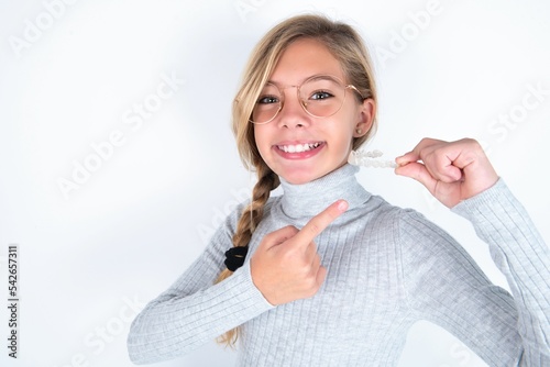 beautiful caucasian teen girl wearing gray turtleneck sweater over white wall holding an invisible aligner and pointing at it. Dental healthcare and confidence concept.