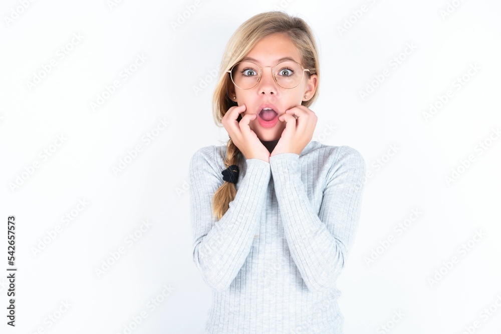 Speechless beautiful caucasian teen girl wearing gray turtleneck sweater over white wall keeps hands near opened mouth reacts to shocking news stares wondered at camera Speechless beautiful caucasian teen girl wearing gray turtleneck sweater over white wall keeps hands near opened mouth reacts to shocking news stares wondered at camera