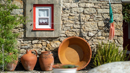 potes de olaria num pátio, encostados, a um muro de casa de pedra, com uma pequena janela com caixilho vermelho e cortina de renda photo