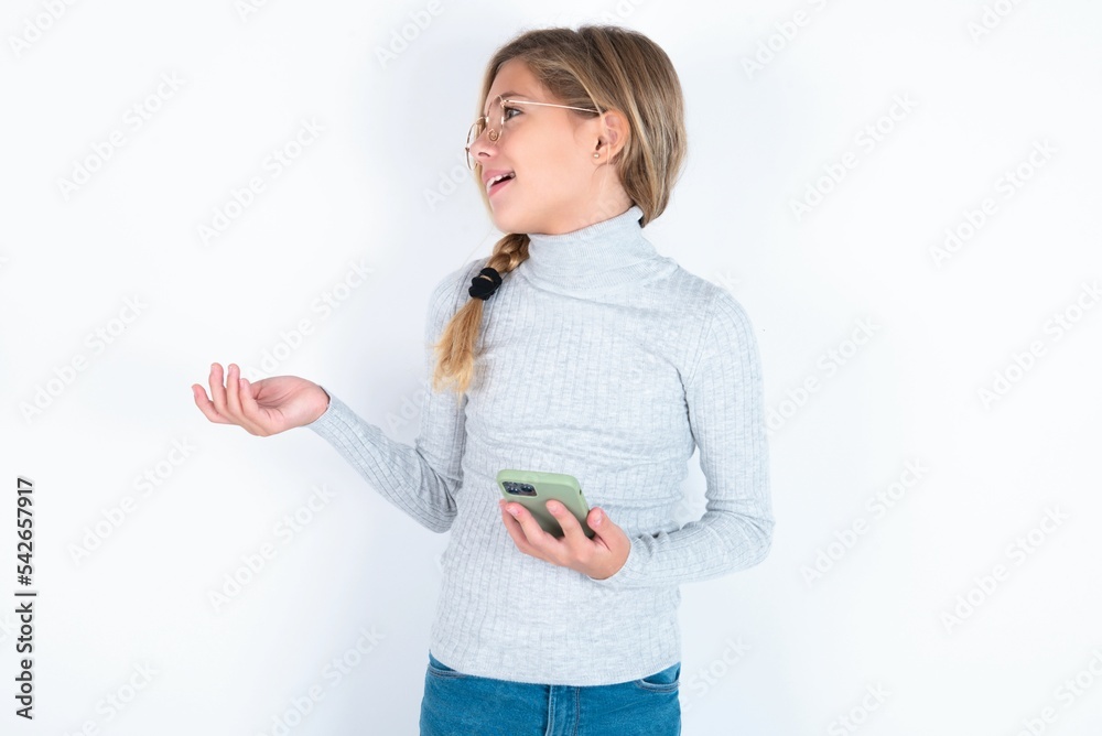 Happy pleased beautiful caucasian teen girl wearing gray turtleneck sweater over white wall raises palm and holds cellphone uses high speed internet for text messaging or video calls Happy pleased beautiful caucasian teen girl wearing gray turtleneck sweater over white wall raises palm and holds cellphone uses high speed internet for text messaging or video calls