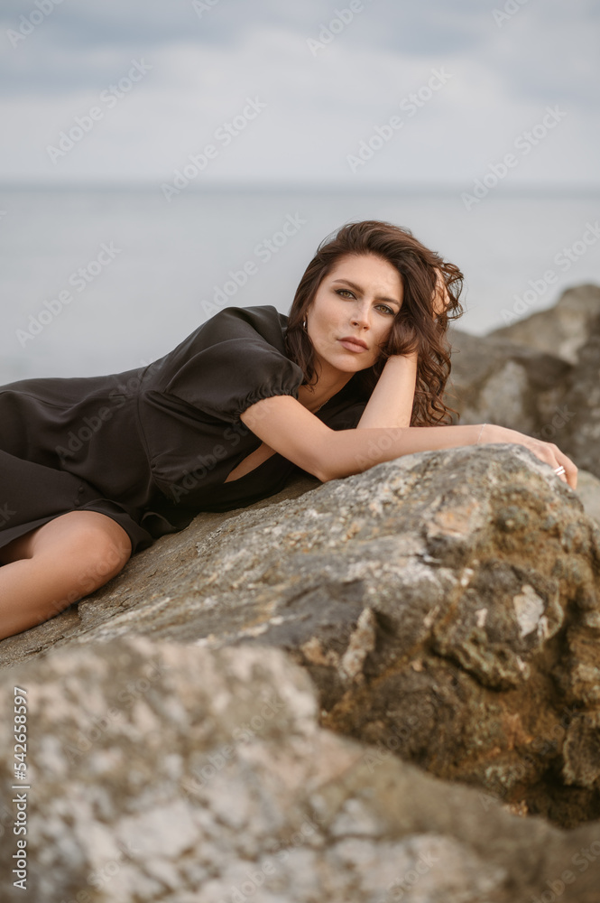  Sexy tanned girl in sits on sea rocks. Portrait of pretty woman dark hair, light eyes.