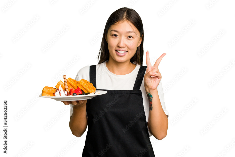 Young asian waitress woman holding waffles isolated joyful and carefree showing a peace symbol with fingers. Young asian waitress woman holding waffles isolated joyful and carefree showing a peace symbol with fingers.