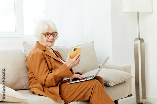 a happy elderly woman in a brown suit is sitting on the sofa and smiling pleasantly looking at her smartphone in a bright case © Tatiana