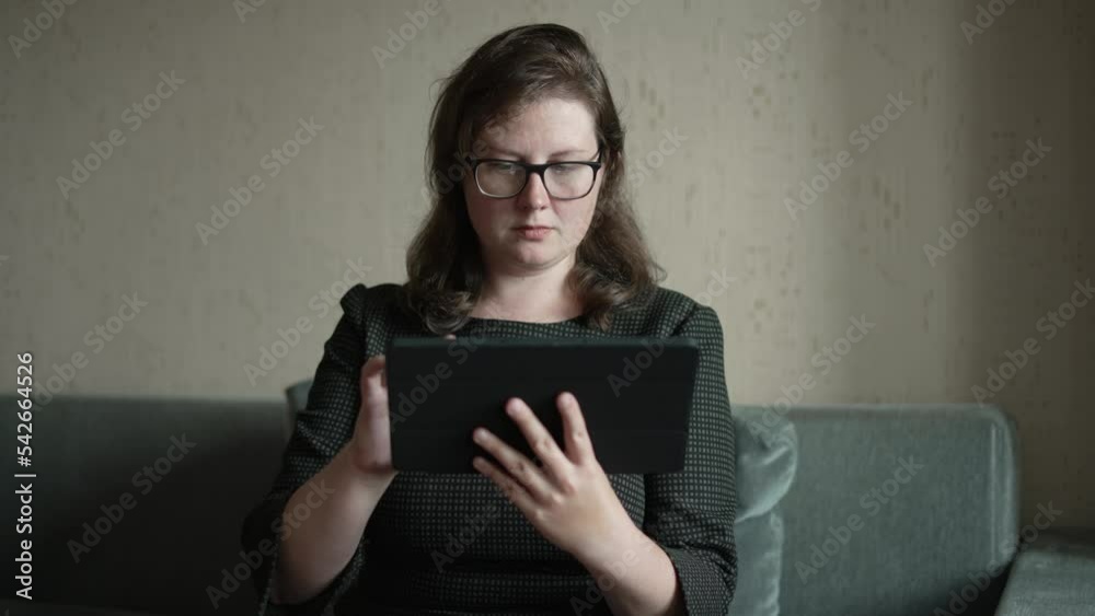 Focused woman in glasses sits, looks attentively at screen of tablet. Focused woman in glasses sits, looks attentively at screen of tablet.