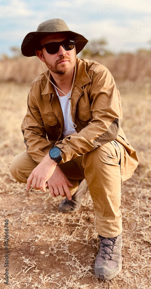 Attractive young man in a safari outfit with a hat and sunglasses crouching in the plain. Safari background