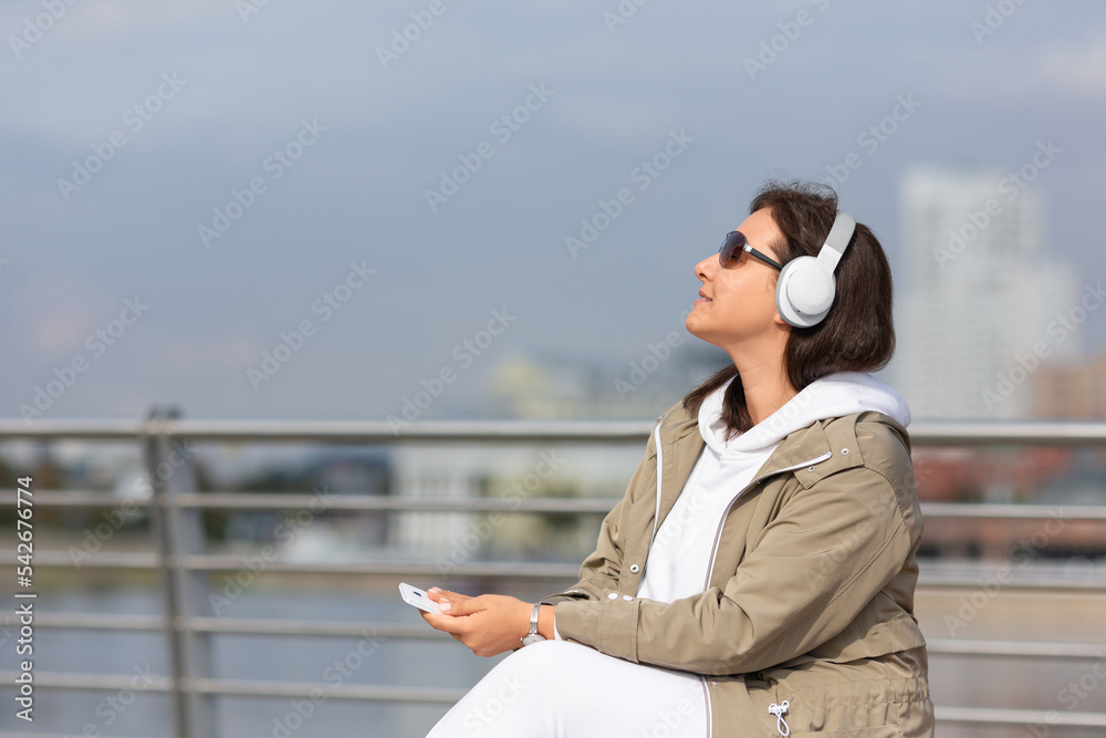 Attractive woman with headphones and mobile in hand sits on the city embankment and enjoying music and privacy, selective focus. Attractive woman with headphones and mobile in hand sits on the city embankment and enjoying music and privacy, selective focus.