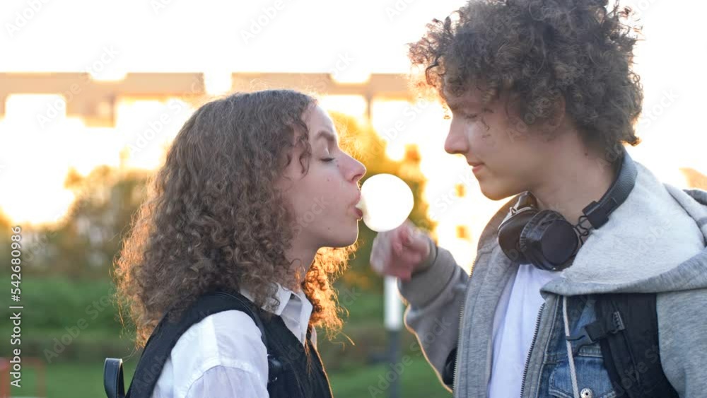 Couple of high school friends outdoors. Girl blowing up a bubble gum ball. The guy doesn't like it very much.