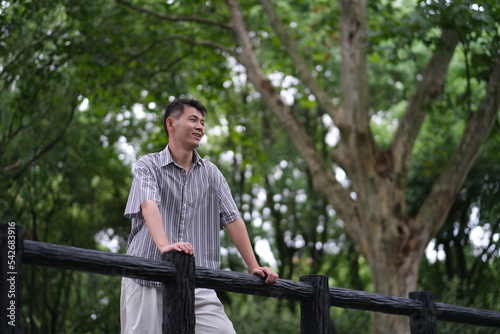 smiling Asian young man at outdoor green forest park at summer, looking away. Low angle
