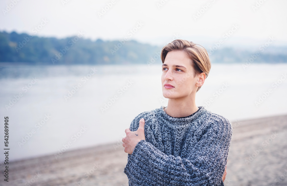 Young stylish blonde woman with a short haircut in a sweater standing on the river bank hugging herself. Androgenic authentic beauty and non-binary diversity personalities Young stylish blonde woman with a short haircut in a sweater standing on the river bank hugging herself. Androgenic authentic beauty and non-binary diversity personalities