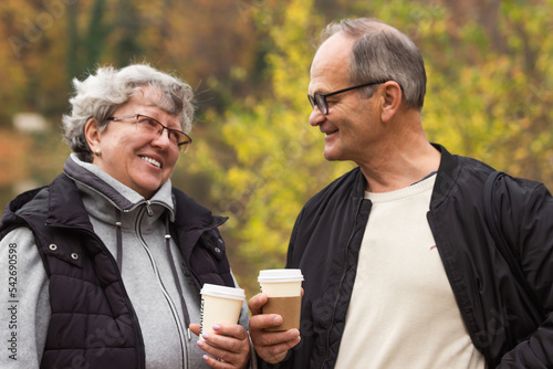 Happy senior couple walking in autumn park and drinking coffee. Elderly couple holding disposable cups, coffee to go.