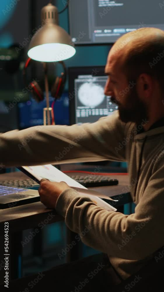 Vertical video: Software developer analyzing web data on clipboard papers and screens, comparing information on files. Young man using programming language and coding new user interface at night. Vertical video: Software developer analyzing web data on clipboard papers and screens, comparing information on files. Young man using programming language and coding new user interface at night.