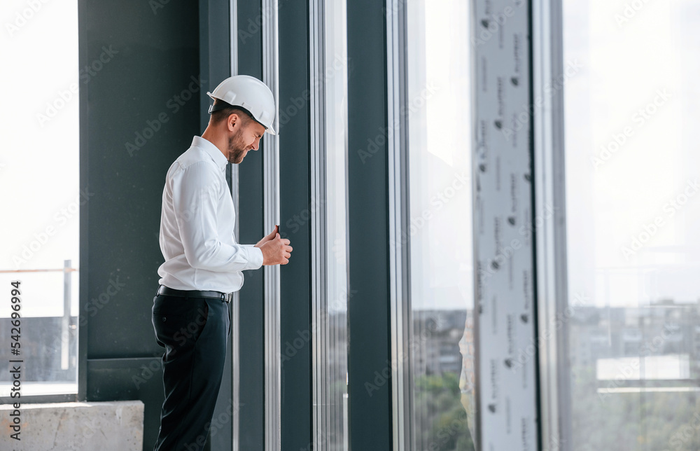 Standing near panoramic windows. Man in white shirt is working indoors at construction site. Managing the project Standing near panoramic windows. Man in white shirt is working indoors at construction site. Managing the project
