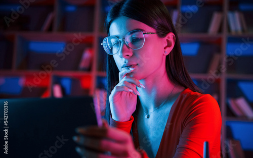 Beautiful woman in glasses and red wear is sitting by the laptop in dark room with neon lighting