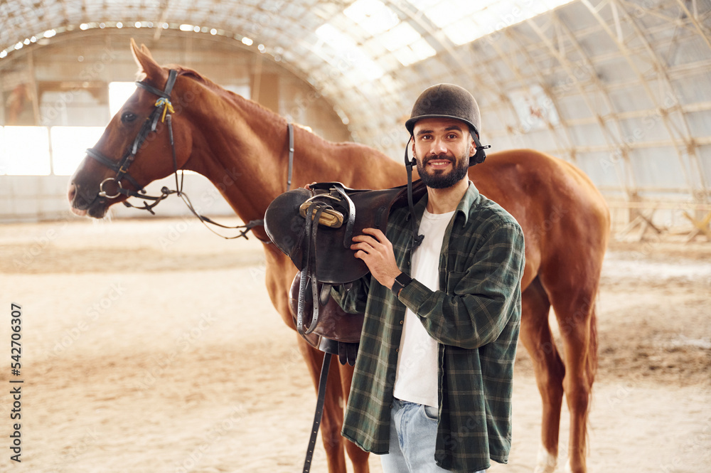 Obraz premium Holding the saddle in hands. Young man with a horse is in the hangar