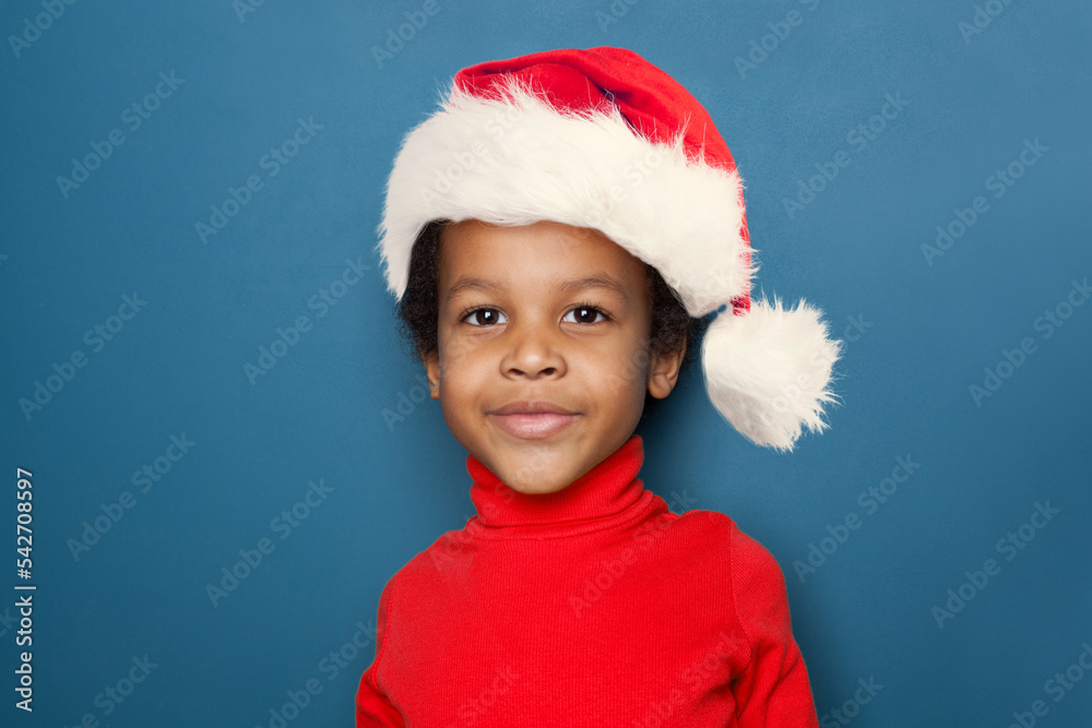Curious black kid portrait. Little child boy in Santa hat smiling on blue background Curious black kid portrait. Little child boy in Santa hat smiling on blue background