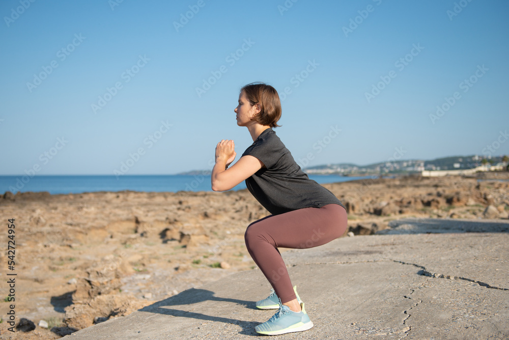 Woman in sportswear doing squat exercise at seaside. Female practicing workout outdoors during sunny day