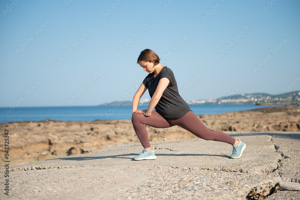 Woman in sportswear doing stretching exercise at seaside. Female practicing workout outdoors during sunny day