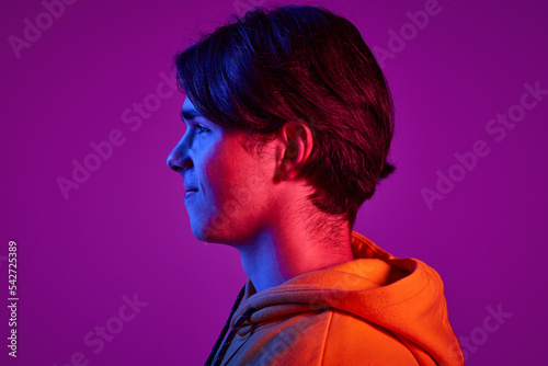 Portrait of young man in casual hoodie posing, looking away isolated over purple background in neon light. Side view.